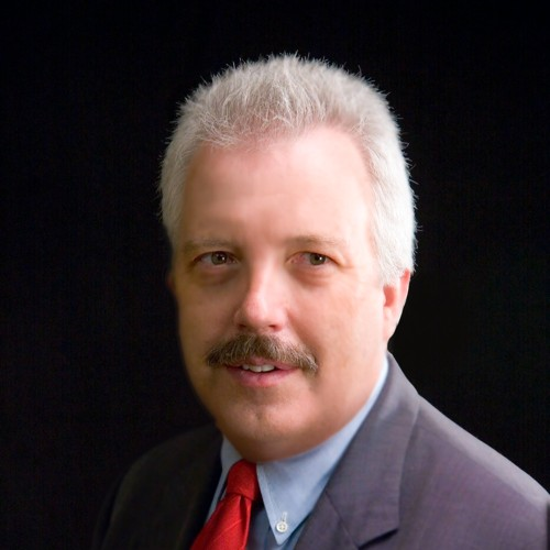 A professional headshot of Dennis Briscoe, an older man with silver hair and a light brown mustache, wearing a grey suit and red tie.